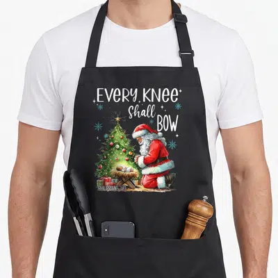 Shall santa apron - Santa kneeling by manger and tree with text Every Knee Shall Bow Philippians 2:10 — shown on a black cotton canvas apron against a white background