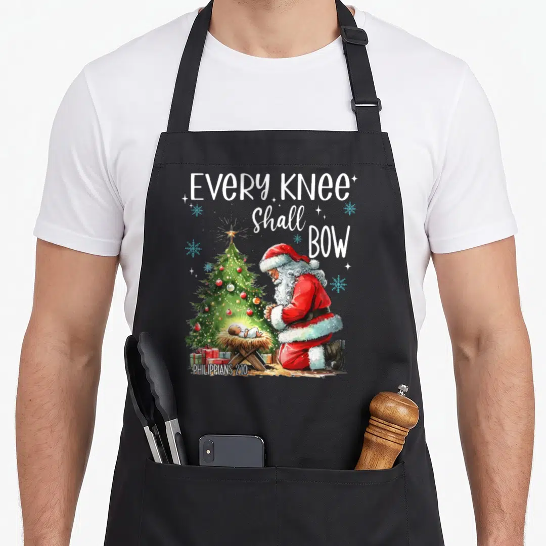 Shall santa apron - Santa kneeling by manger and tree with text Every Knee Shall Bow Philippians 2:10 — shown on a black cotton canvas apron against a white background
