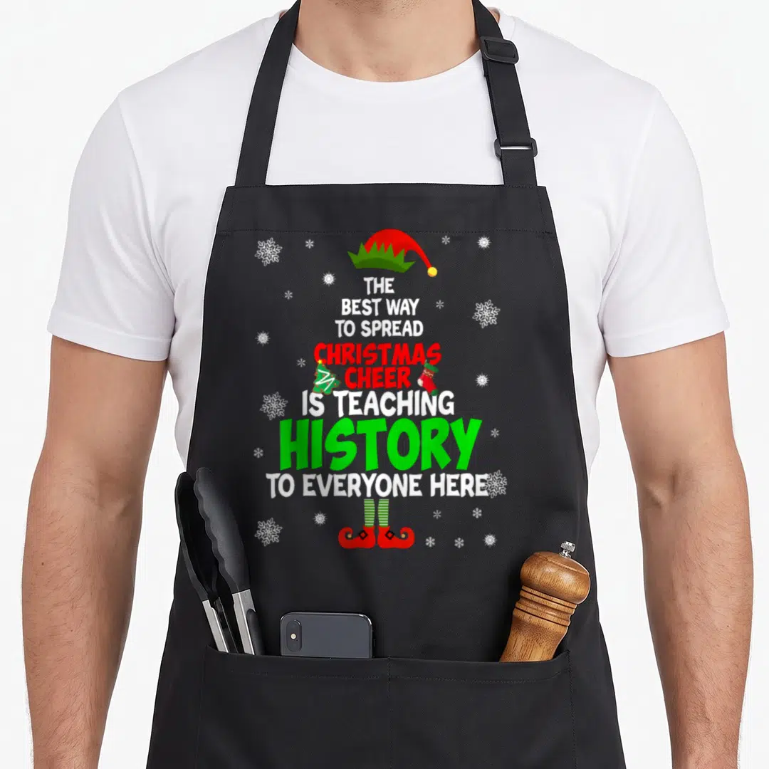 Spread christmas apron - Stacked text reading teaching history surrounded by elf hat, shoes, tree, stocking — shown on a black cotton canvas apron against a white background