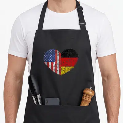 Dual germany apron - Heart split into US and German flag sections with distressed red white blue black gold — shown on a black cotton canvas apron against a white background