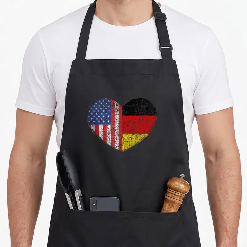 Dual germany apron - Heart split into US and German flag sections with distressed red white blue black gold — shown on a black cotton canvas apron against a white background
