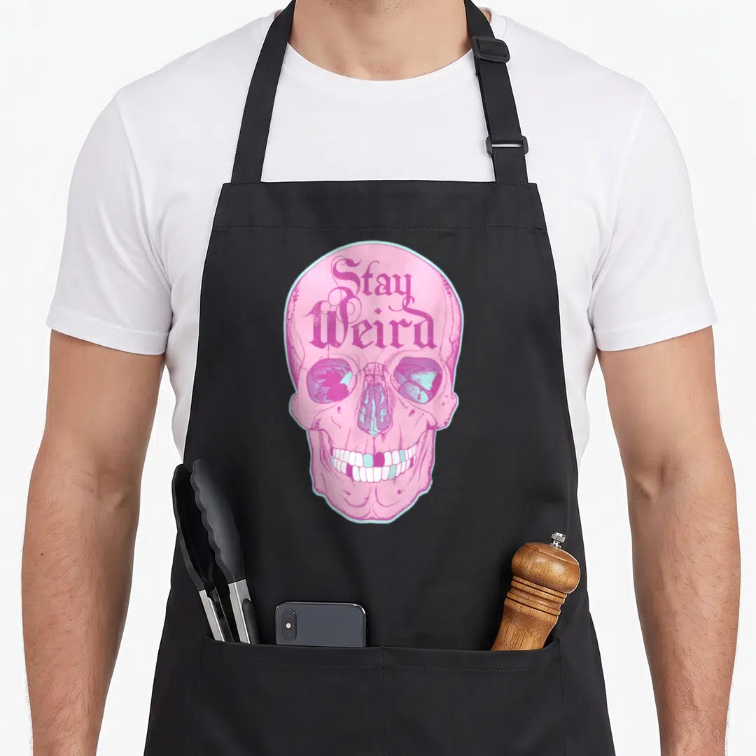 Goth pastel pink apron - Pink skull with light blue outline and Stay Weird text on forehead — shown on a black cotton canvas apron against a white background
