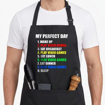 Perfect gaming apron - Typographic list MY PERFECT DAY with colored text items like PLAY VIDEO GAMES in red, yellow — shown on a black cotton canvas apron against a white background
