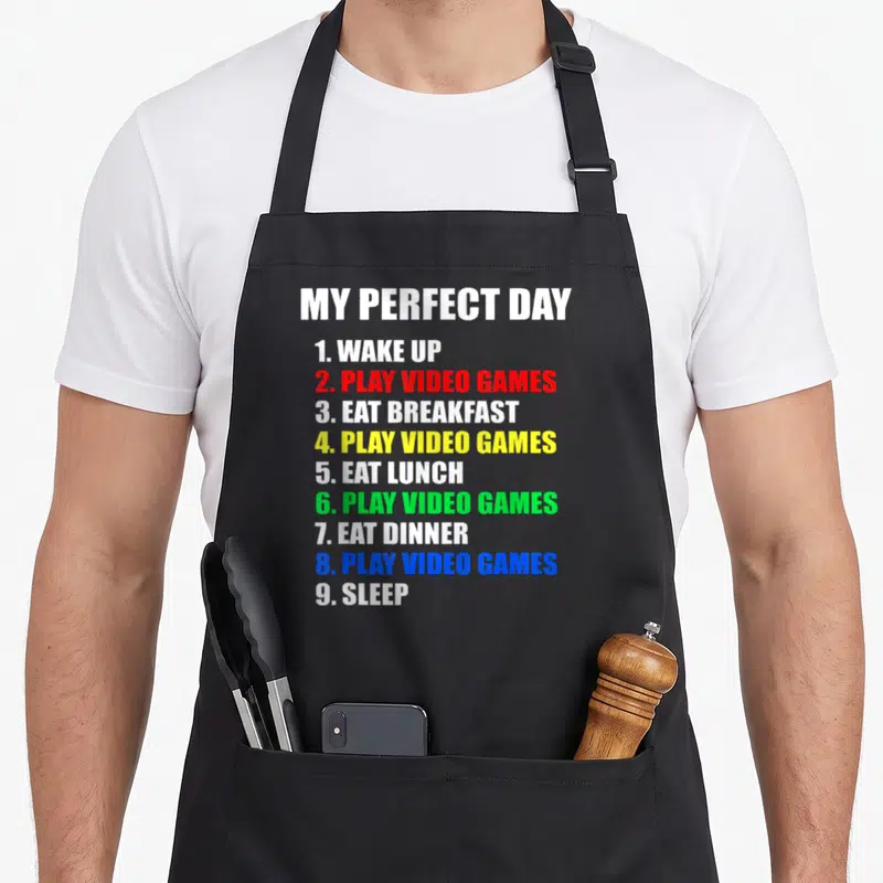 Perfect gaming apron - Typographic list MY PERFECT DAY with colored text items like PLAY VIDEO GAMES in red, yellow — shown on a black cotton canvas apron against a white background