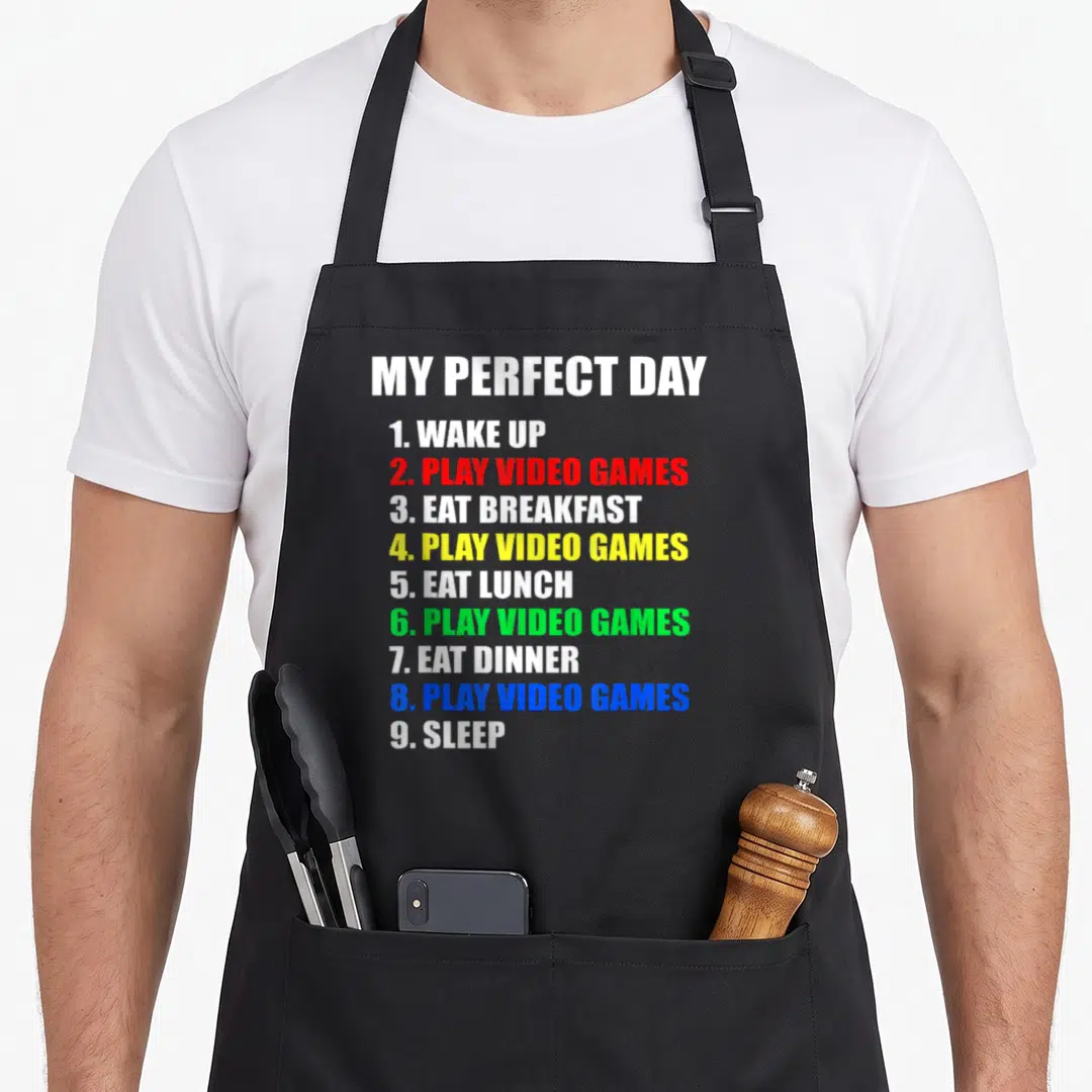Perfect gaming apron - Typographic list MY PERFECT DAY with colored text items like PLAY VIDEO GAMES in red, yellow — shown on a black cotton canvas apron against a white background