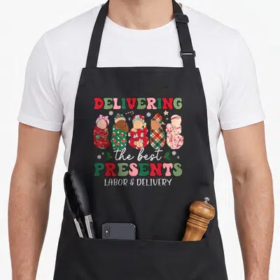 Delivering christmas apron - Five swaddled babies with DELIVERING the best PRESENTS LABOR & DELIVERY text in red green — shown on a black cotton canvas apron against a white background