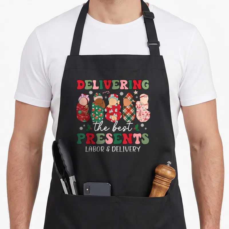 Delivering christmas apron - Five swaddled babies with DELIVERING the best PRESENTS LABOR & DELIVERY text in red green — shown on a black cotton canvas apron against a white background