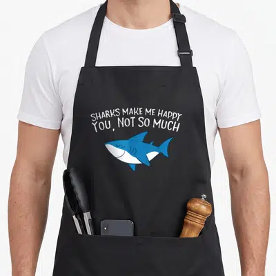 Shark much apron - Blue cartoon shark swimming under white text reading Sharks Make Me Happy You Not So Much — shown on a black cotton canvas apron against a white background