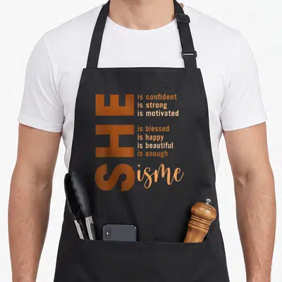 She african american apron - Vertical orange SHE text with stacked affirmations and cursive is me with a sparkle icon — shown on a black cotton canvas apron against a white background