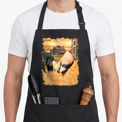 Enthusiast rooster apron - Photo-style rooster standing in grass with barn background and vertical ROOSTER banner — shown on a black cotton canvas apron against a white background