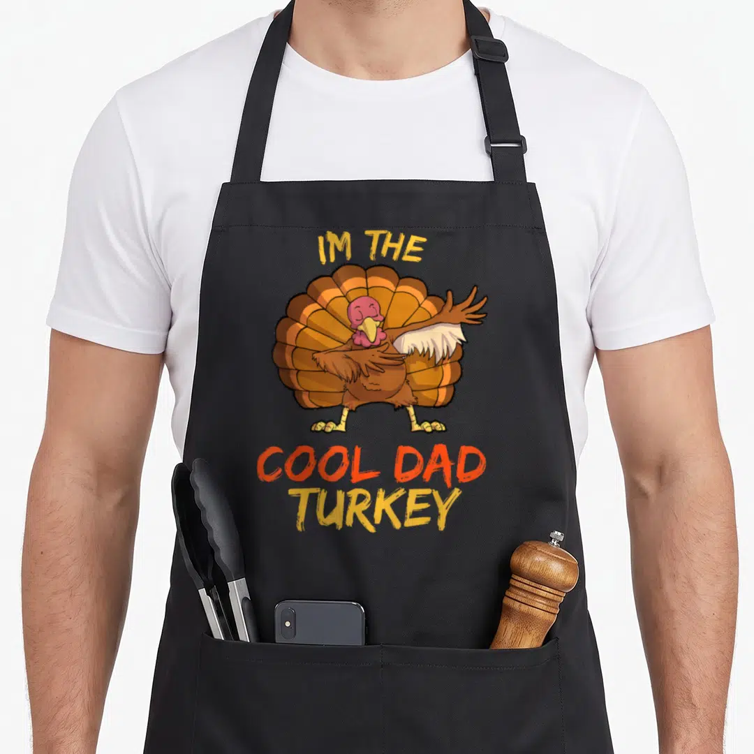 Funny turkey family thanksgiving apron - Cartoon turkey with IM THE COOL DAD TURKEY text in orange and yellow — shown on a black cotton canvas apron against a white background