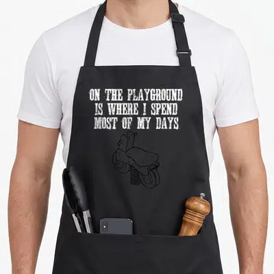Funny most playground apron - White stacked text reading ON THE PLAYGROUND above a black line-art spring scooter — shown on a black cotton canvas apron against a white background