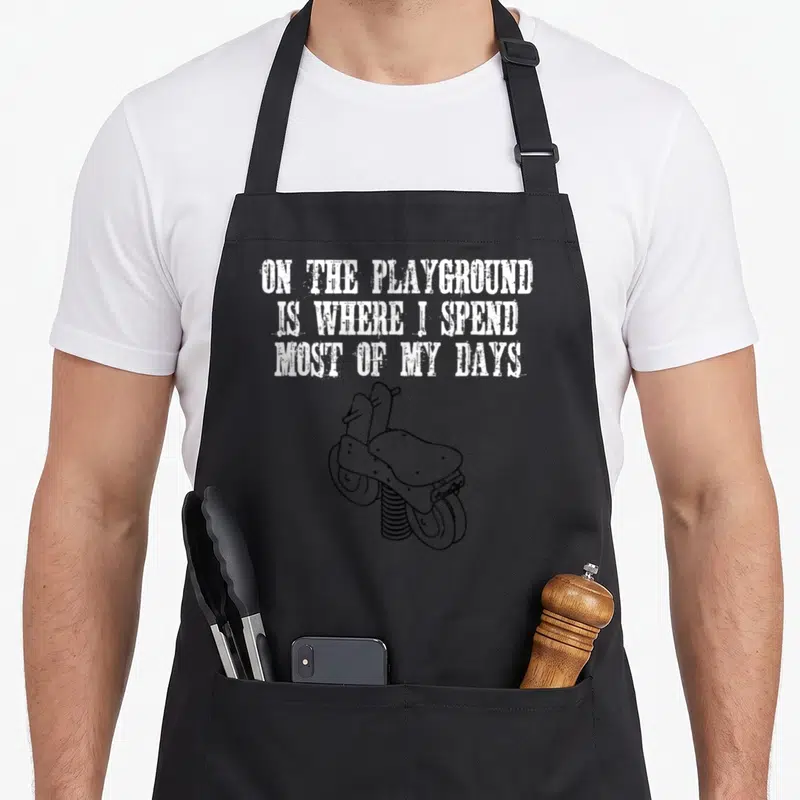 Funny most playground apron - White stacked text reading ON THE PLAYGROUND above a black line-art spring scooter — shown on a black cotton canvas apron against a white background
