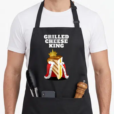 Grilled king apron - Cartoon grilled cheese sandwich with gold crown and red white cape under stacked white text — shown on a black cotton canvas apron against a white background