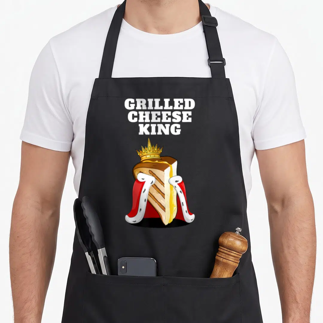 Grilled king apron - Cartoon grilled cheese sandwich with gold crown and red white cape under stacked white text — shown on a black cotton canvas apron against a white background