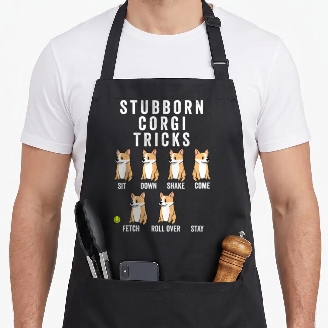 Pembroke corgi aprons - Six tan white corgis sitting under STUBBORN CORGI TRICKS text with tennis ball — shown on a black cotton canvas apron against a white background