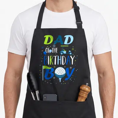 Birthday golf apron - DAD OF THE BIRTHDAY BOY text with golf cart, ball, flag, and confetti in blue and green — shown on a black cotton canvas apron against a white background