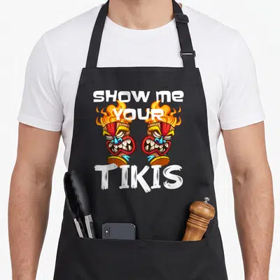 Show spring aprons - Two cartoon tiki torches with fire and text SHOW ME YOUR TIKIS in white, orange, red, and brown — shown on a black cotton canvas apron against a white background