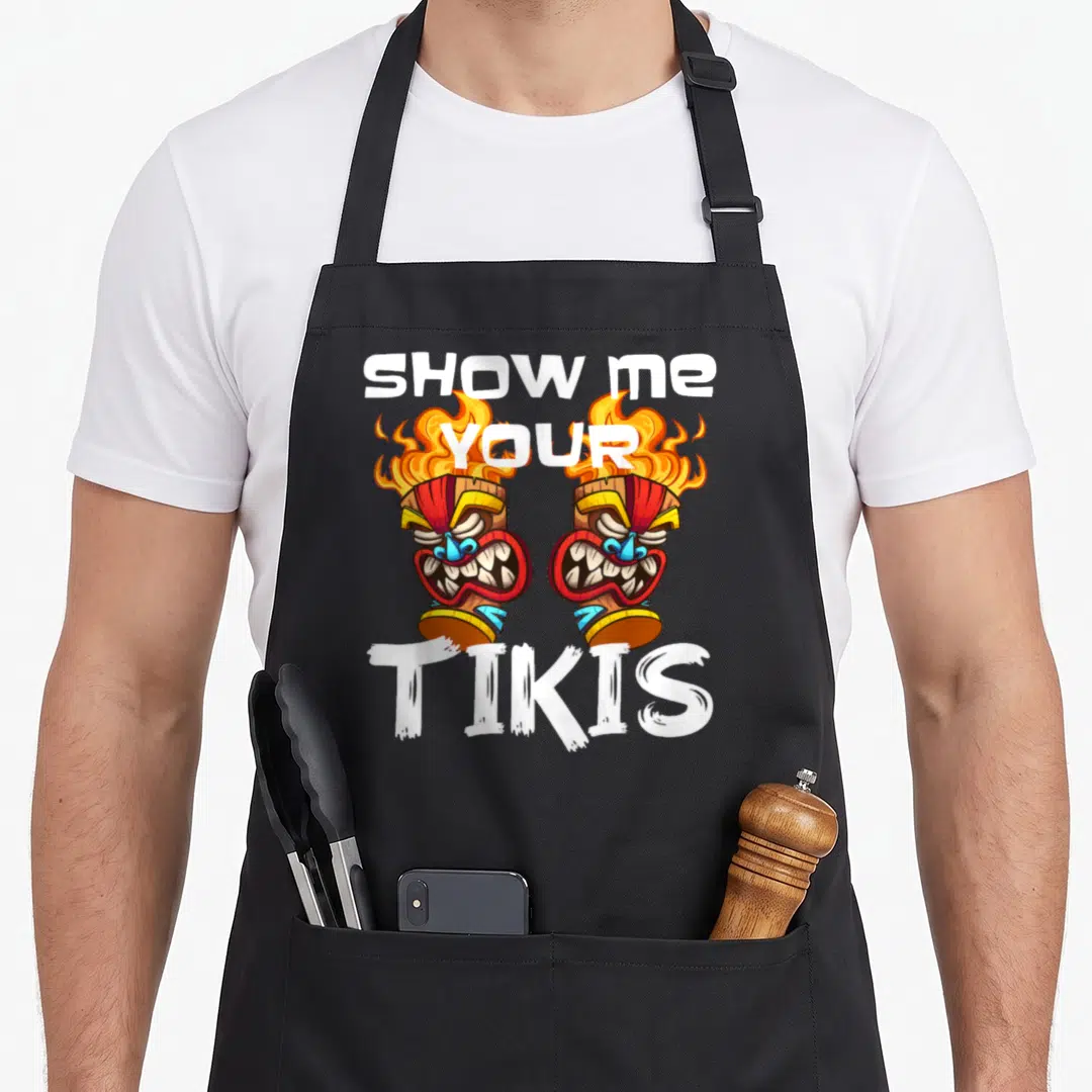 Show spring aprons - Two cartoon tiki torches with fire and text SHOW ME YOUR TIKIS in white, orange, red, and brown — shown on a black cotton canvas apron against a white background