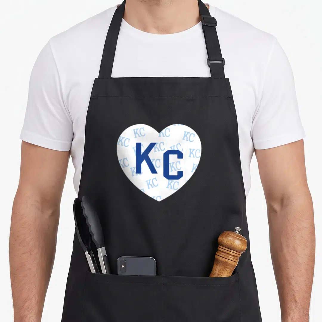 Pattern kansas girl apron - White heart with large blue KC and scattered light blue KC letters — shown on a black cotton canvas apron against a white background
