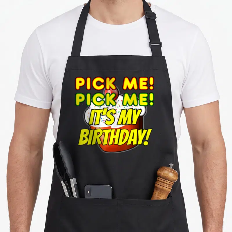 Pick cake apron - PICK ME IT'S MY BIRTHDAY text over yellow and red cartoon cupcake with cherry — shown on a black cotton canvas apron against a white background