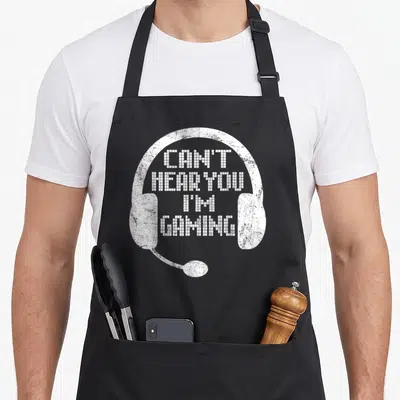 Headset hear gaming apron - White headset graphic with pixelated text reading CAN'T HEAR YOU I'M GAMING — shown on a black cotton canvas apron against a white background