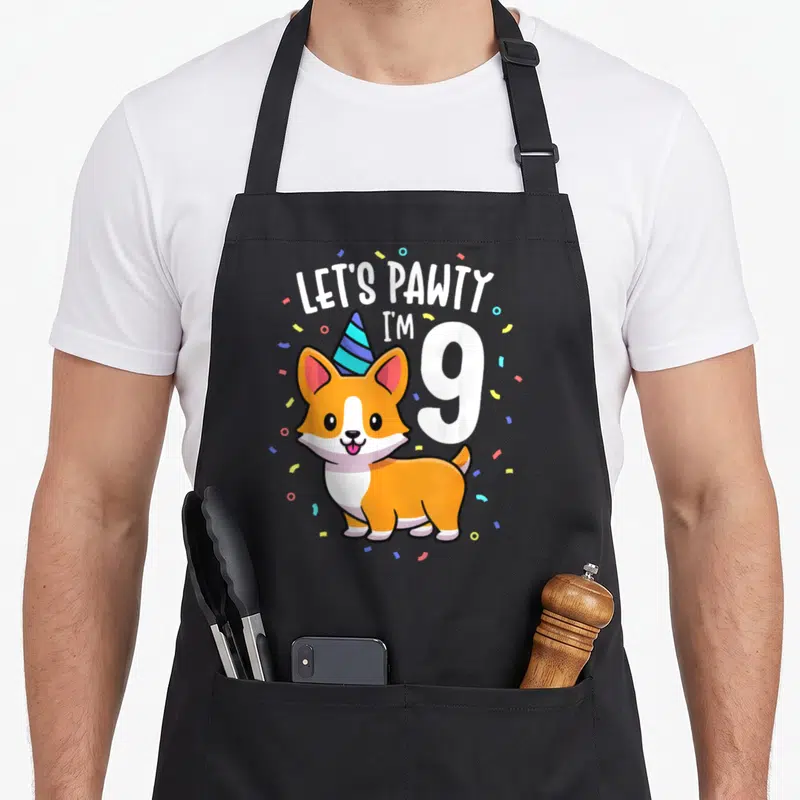 9th corgi apron - Cartoon corgi with party hat and text LET'S PAWTY I'M 9 with confetti — shown on a black cotton canvas apron against a white background