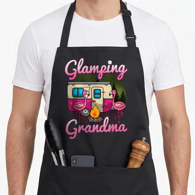 Camper grandma apron - Pink camper with two flamingos and campfire under Glamping Grandma text — shown on a black cotton canvas apron against a white background