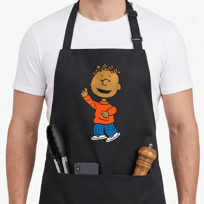 Armstrong peanut apron - Cartoon boy with brown skin in orange shirt and blue striped pants waving — shown on a black cotton canvas apron against a white background