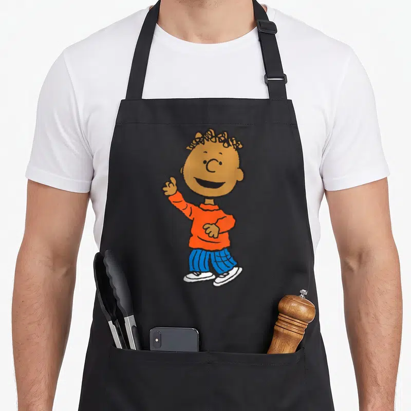Armstrong peanut apron - Cartoon boy with brown skin in orange shirt and blue striped pants waving — shown on a black cotton canvas apron against a white background