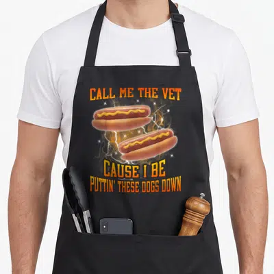 Call hot dog apron - Two hot dogs with lightning bolts and CALL ME THE VET text in orange and yellow — shown on a black cotton canvas apron against a white background