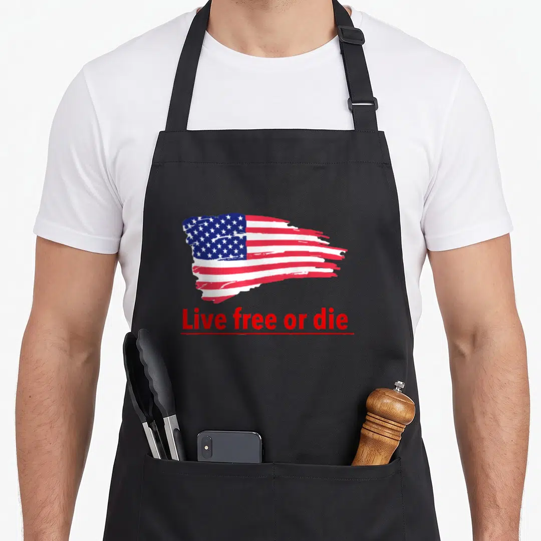 Live patriotic apron - Brush-stroke US flag above red text Live free or die with underline — shown on a black cotton canvas apron against a white background