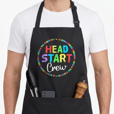 Education preschool teacher apron - Colorful stacked HEAD START Crew text surrounded by circular border of kindness — shown on a black cotton canvas apron against a white background