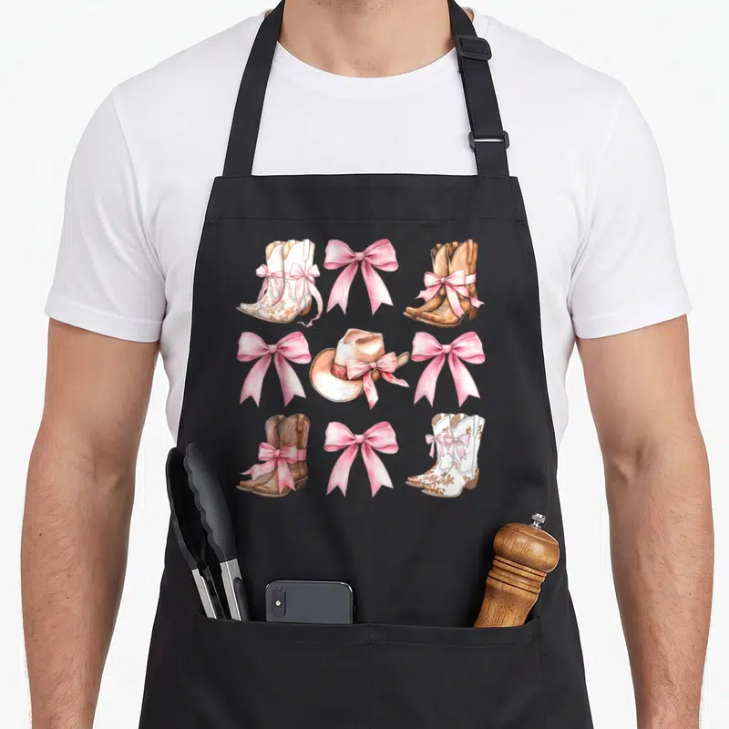 Coquette cowgirl aprons - Grid of white and brown cowboy boots with pink bows and a beige hat — shown on a black cotton canvas apron against a white background
