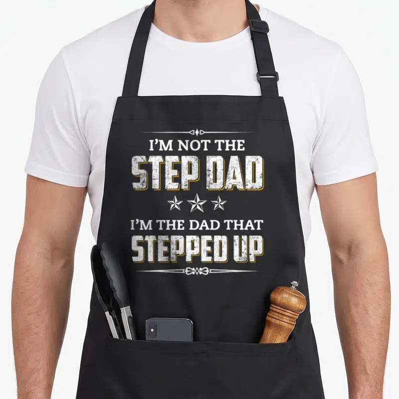 Step stepfather apron - Distressed white and yellow text reading I'M NOT THE STEP DAD with three stars and decorative — shown on a black cotton canvas apron against a white background