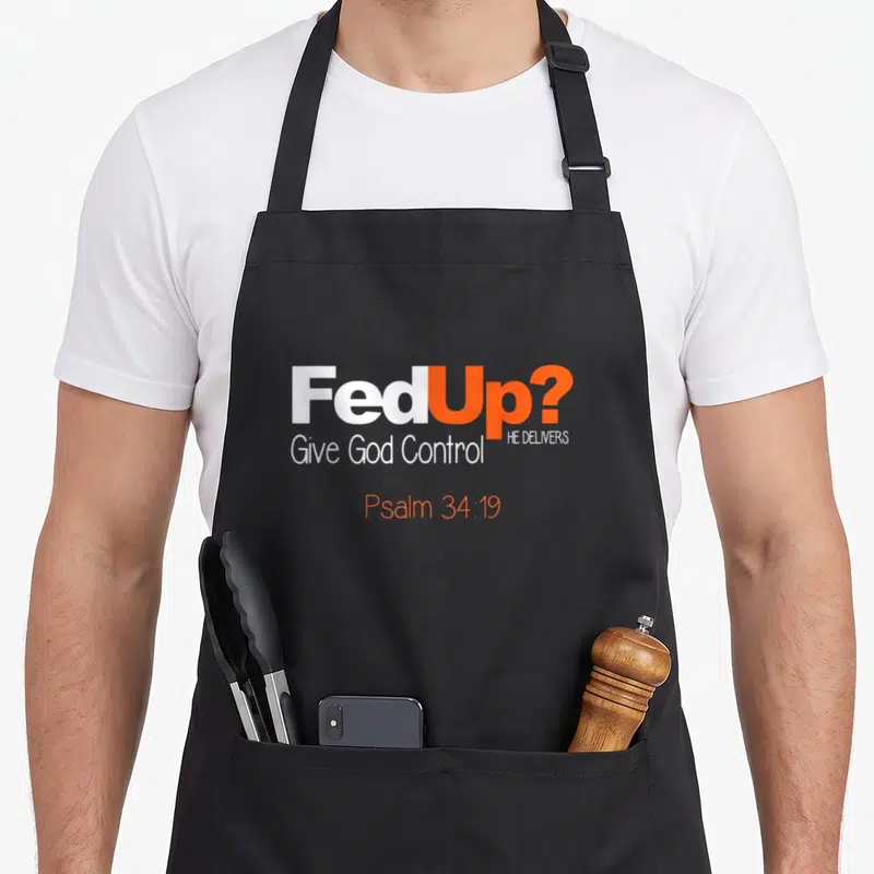 Fed christian apron - FedUp typography with Give God Control and Psalm 34:19 in white and orange text — shown on a black cotton canvas apron against a white background