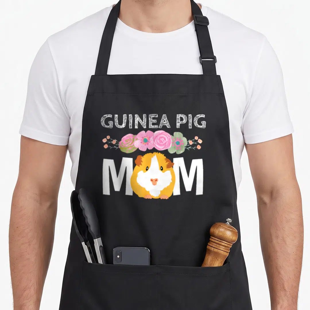 Guinea floral apron - Cartoon guinea pig replacing O in MOM with pink and green flowers — shown on a black cotton canvas apron against a white background
