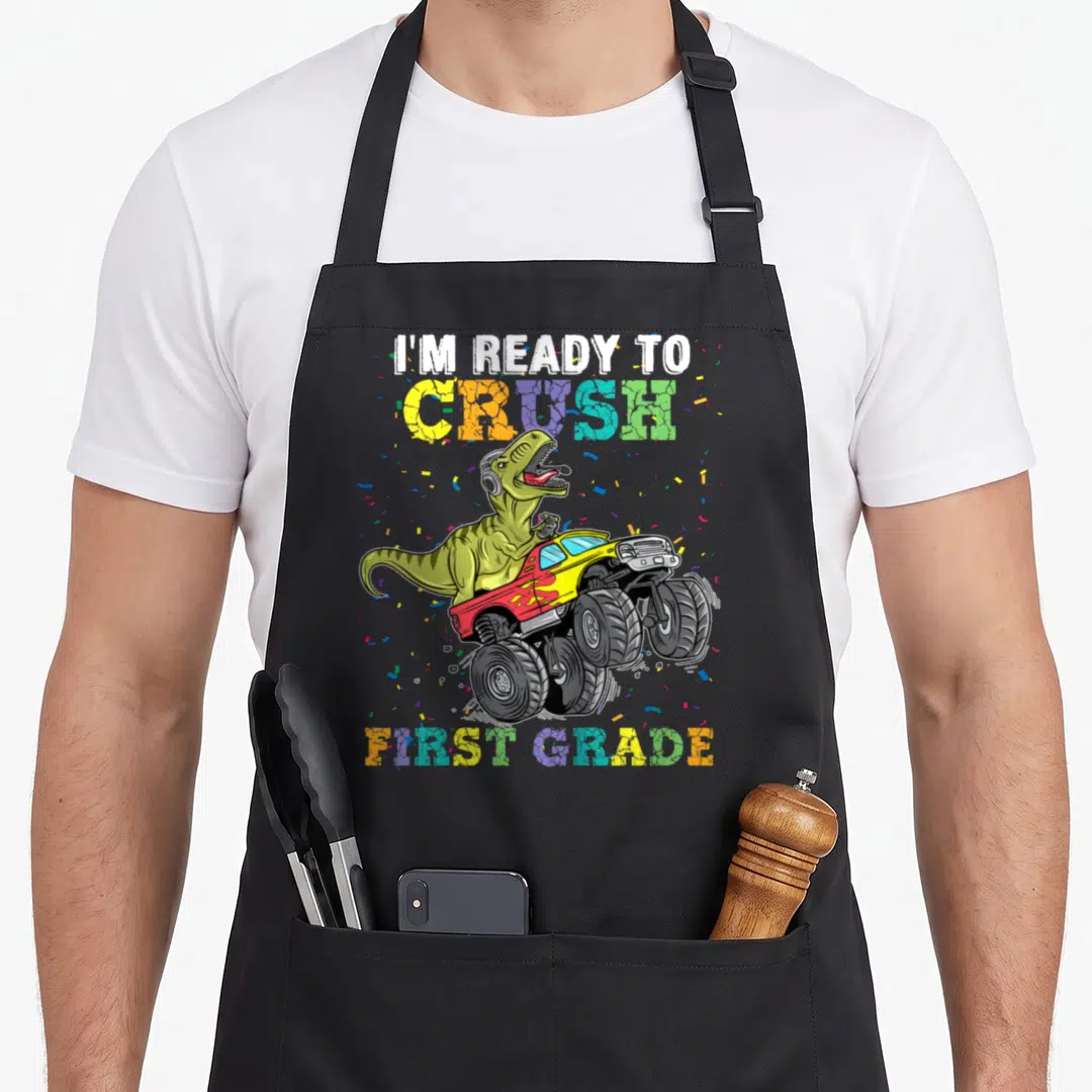 Grade truck apron - T-Rex driving monster truck with I'm Ready to Crush First Grade text and confetti — shown on a black cotton canvas apron against a white background
