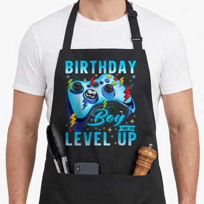 Birthday level video game apron - Blue game controller with lightning bolts and BIRTHDAY Boy TIME TO LEVEL UP text — shown on a black cotton canvas apron against a white background