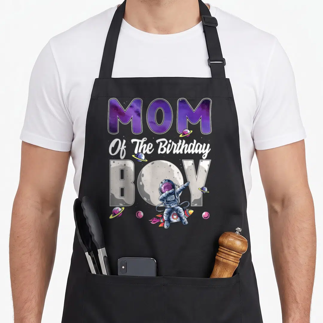 Space theme birthday apron - MOM Of The Birthday BOY text with purple galaxy font and cartoon astronaut dabbing — shown on a black cotton canvas apron against a white background