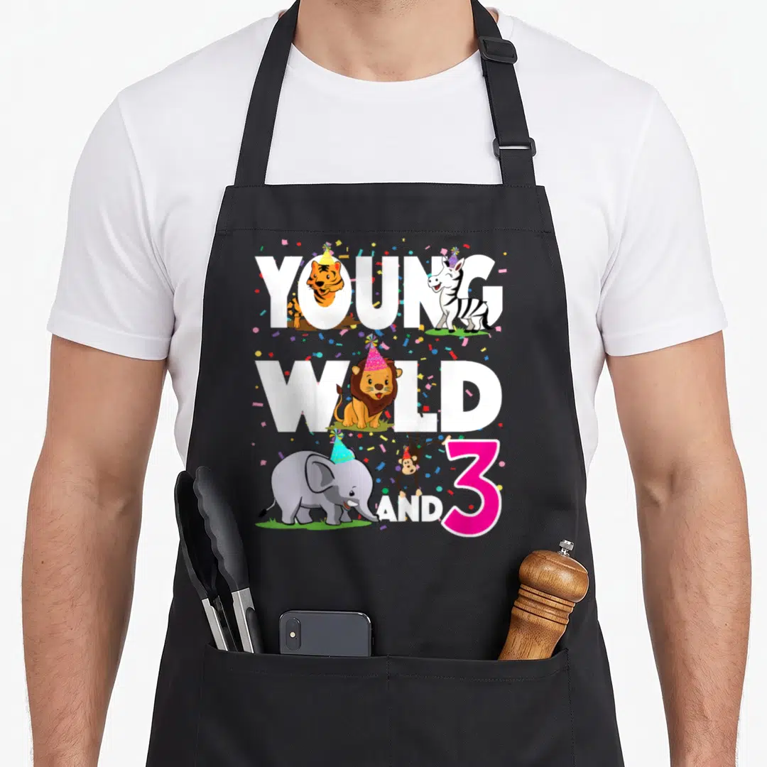 Young birthday apron - Young Wild And 3 text with cartoon animals wearing party hats and confetti — shown on a black cotton canvas apron against a white background