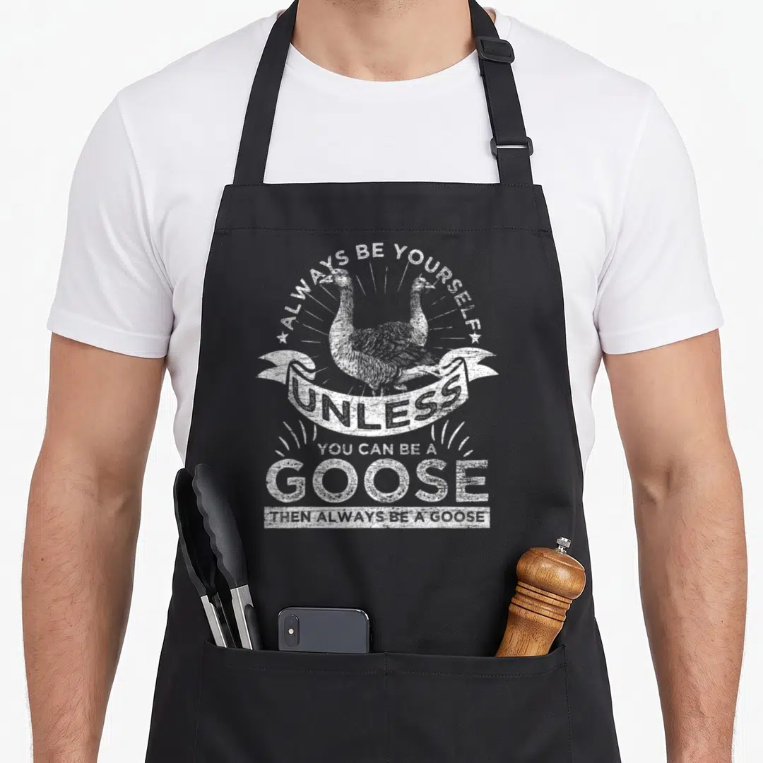 Alway goose apron - White distressed goose illustration with arched text and ribbon banner reading Always Be Yourself — shown on a black cotton canvas apron against a white background