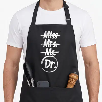 Phd mrs apron - White typography listing Miss Mrs Ms Dr with first three crossed out and Dr circled — shown on a black cotton canvas apron against a white background