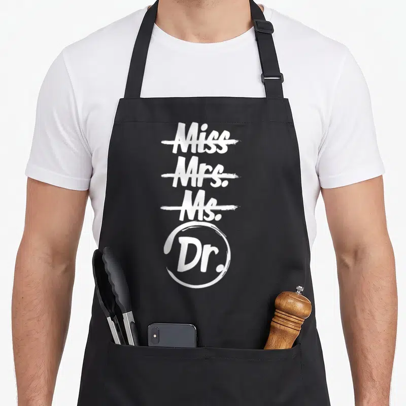 Phd mrs apron - White typography listing Miss Mrs Ms Dr with first three crossed out and Dr circled — shown on a black cotton canvas apron against a white background