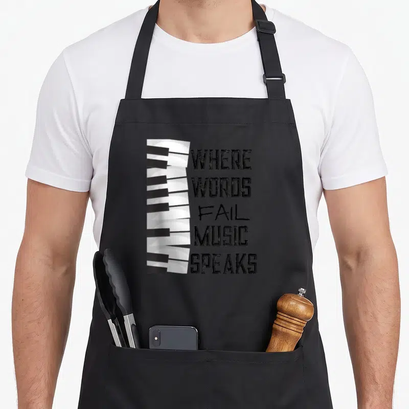 Music themed aprons - Vertical piano keys beside stacked text reading WHERE WORDS FAIL MUSIC SPEAKS — shown on a black cotton canvas apron against a white background