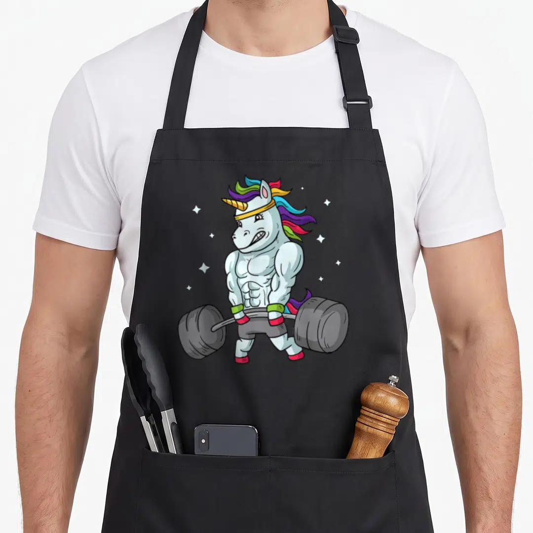 Deadlift unicorn apron - Muscular cartoon unicorn lifting barbell with rainbow mane and white stars on grey background — shown on a black cotton canvas apron against a white background