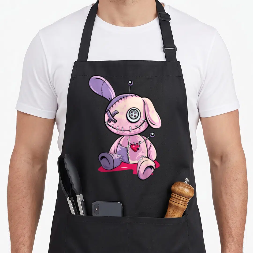 Doll goth apron - Stitched pink rabbit with button eye, pins, and red heart patch in cartoon style — shown on a black cotton canvas apron against a white background