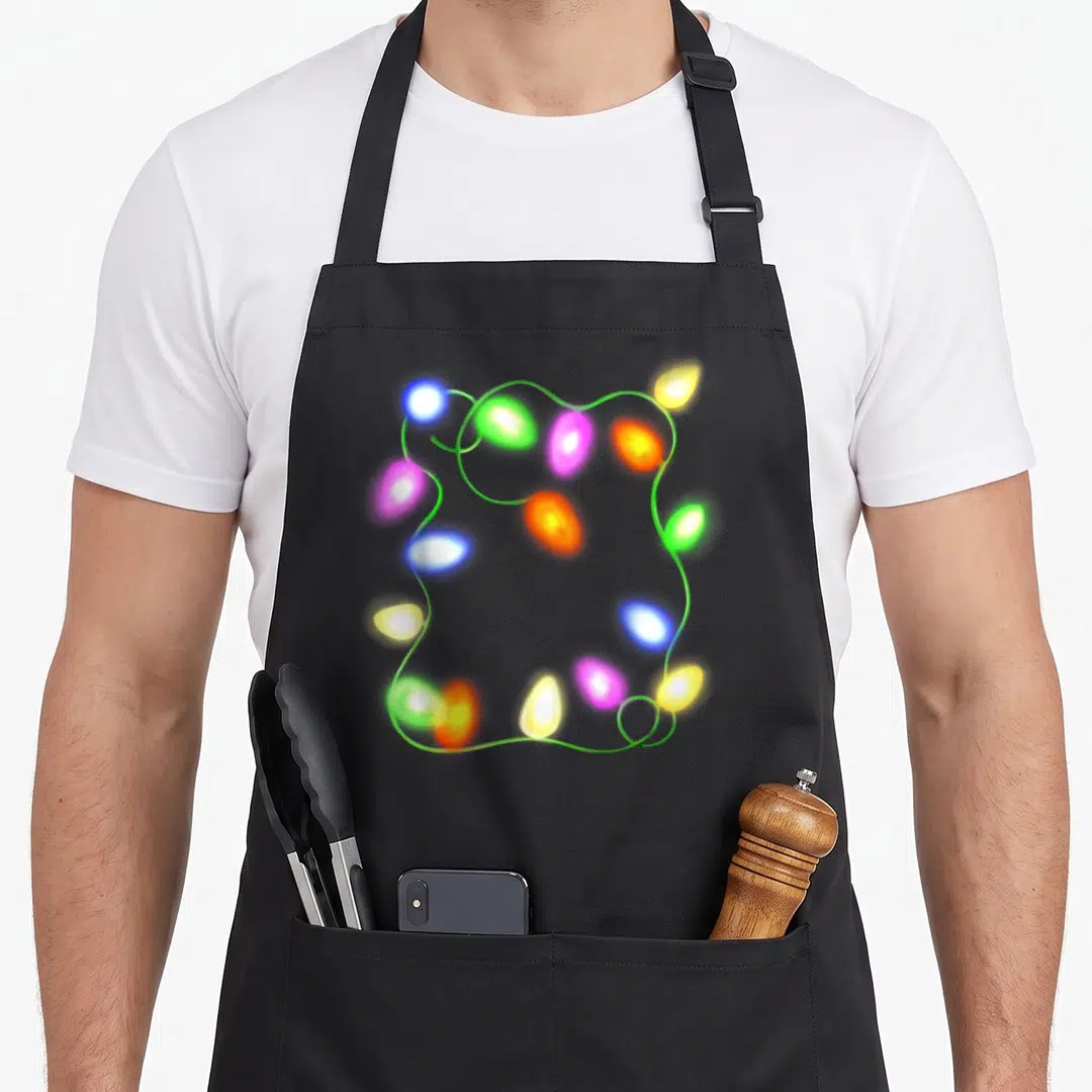 Glowing christmas tree apron - Realistic green wire with glowing green blue yellow pink and orange bulbs in a loose — shown on a black cotton canvas apron against a white background