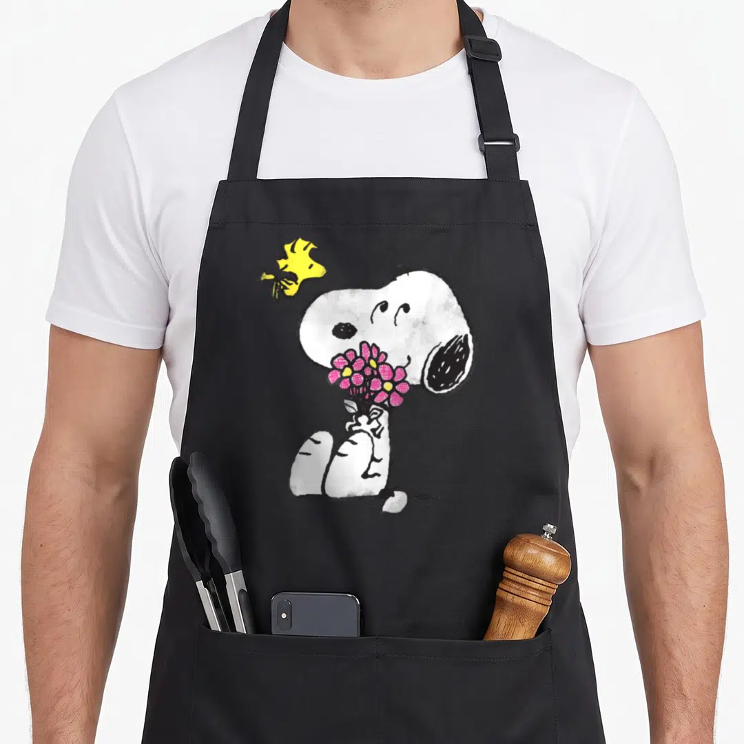 Flower mother's apron - Snoopy sitting holding pink and yellow flowers with Woodstock flying above in cartoon style — shown on a black cotton canvas apron against a white background