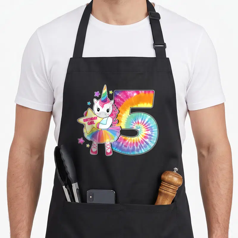 Unicorn tie-dye apron - Cartoon unicorn in tutu holding BIRTHDAY GIRL star beside tie-dye number 5 — shown on a black cotton canvas apron against a white background